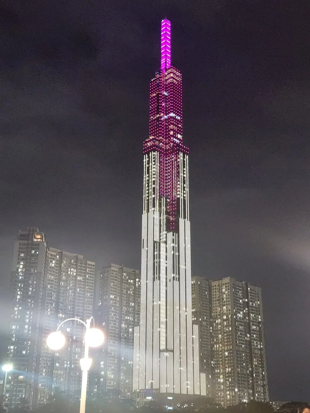 Tall illuminated skyscraper above neighbouring buildings at night in Ho Chi Minh City.